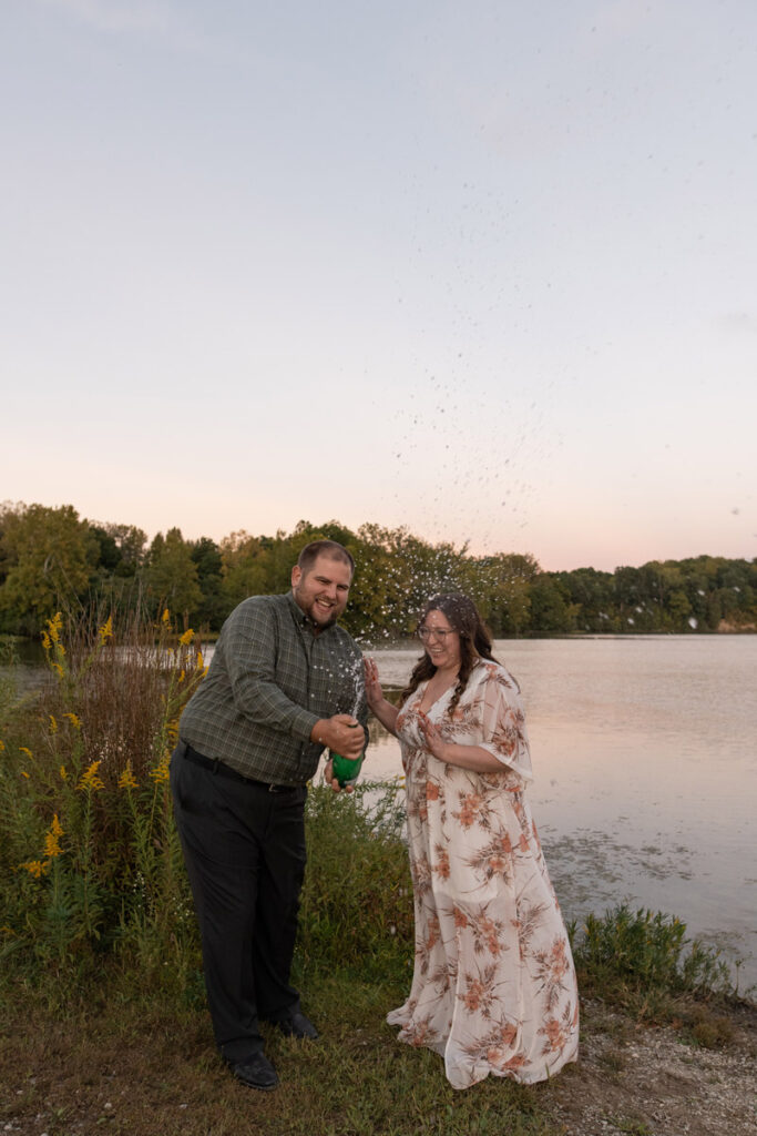 Couple do a sparkling water spray outside at sunset beside a lake.