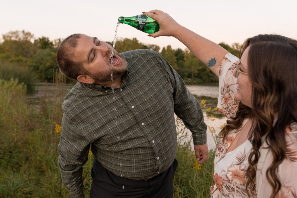 Woman pours sparking water out of glass bottle and man tries to drink it.