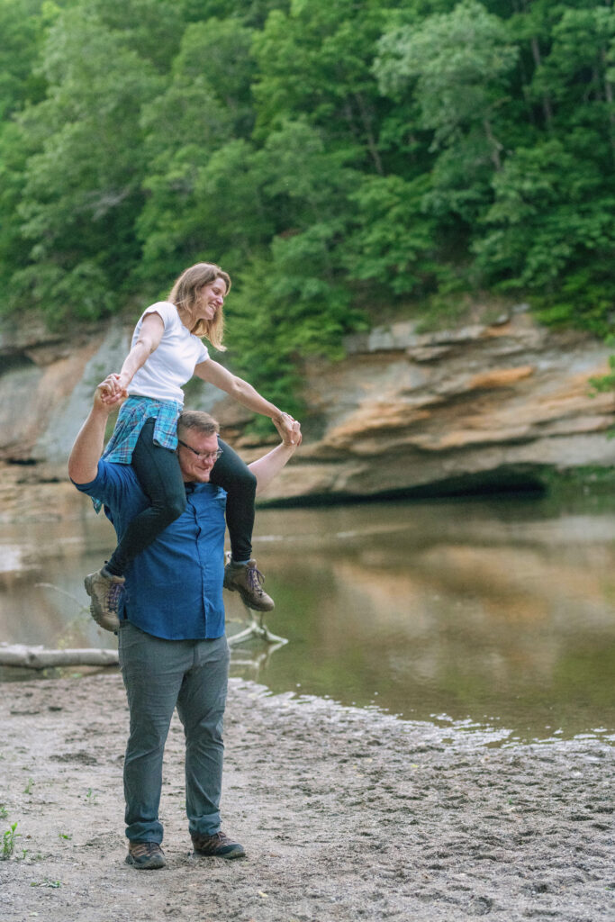 Girl sits on man's shoulders as they walk along a creek shore.