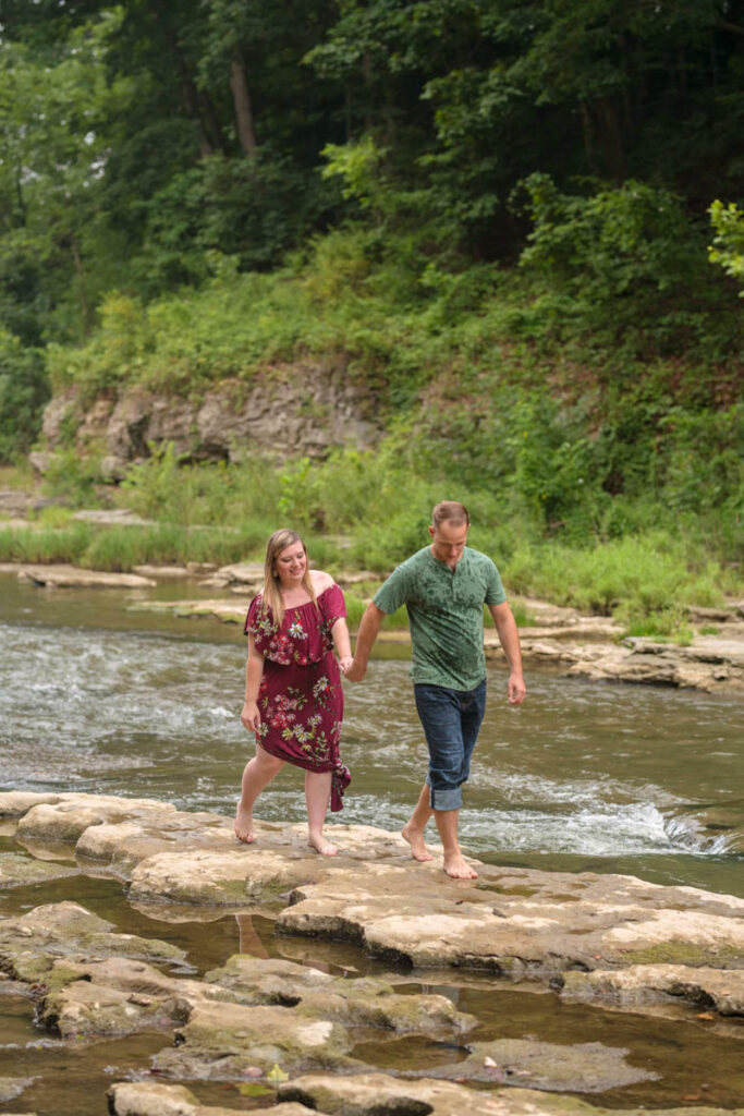 Man leads woman on rocks through water during waterfall engagement photos.