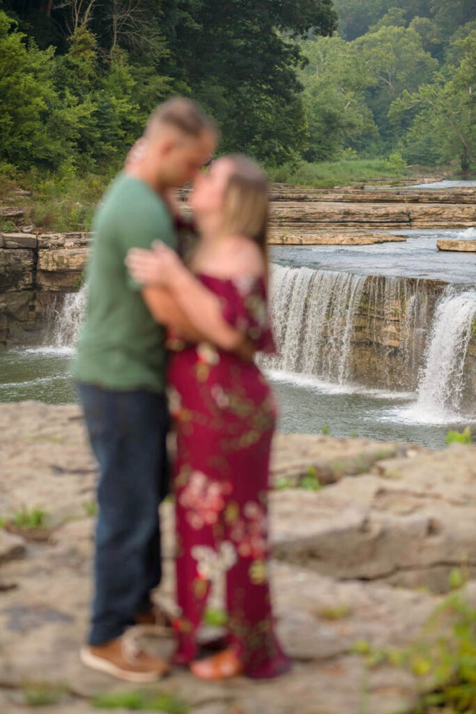 Couple hold one another closely with large waterfalls flowing behind them for waterfall engagement photos at Cataract Falls.
