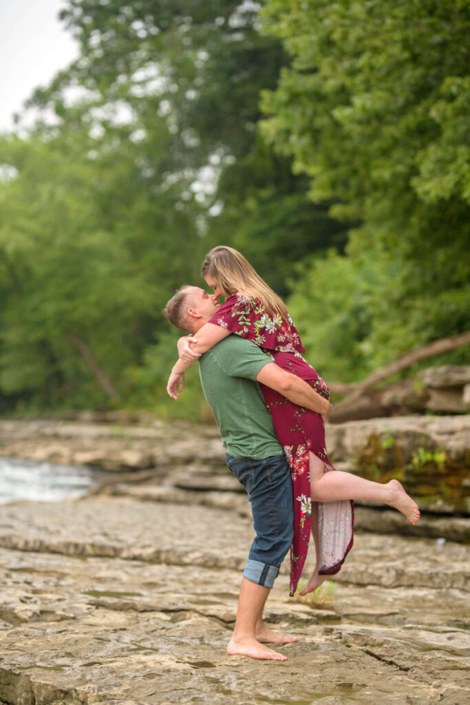 Man lifts up woman during waterfall engagement photos.