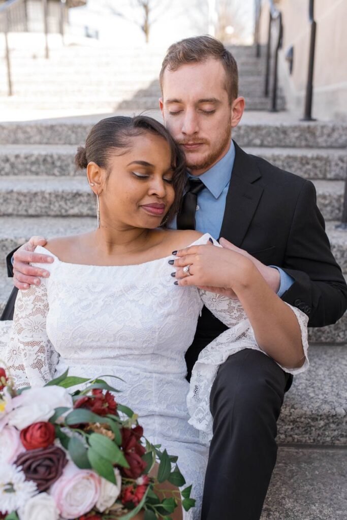 Bride and groom sit closely together with closed eyes on marble steps.