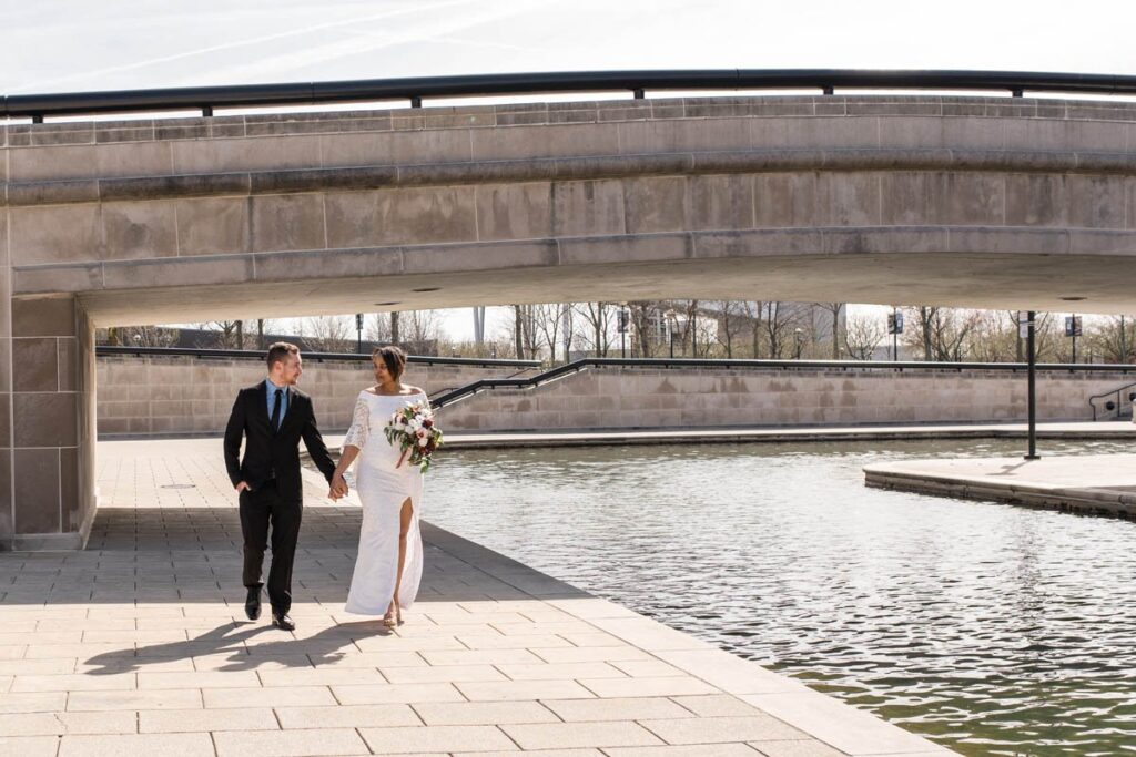 Bride and groom hold hands as they walk along the canal in Indianapolis.