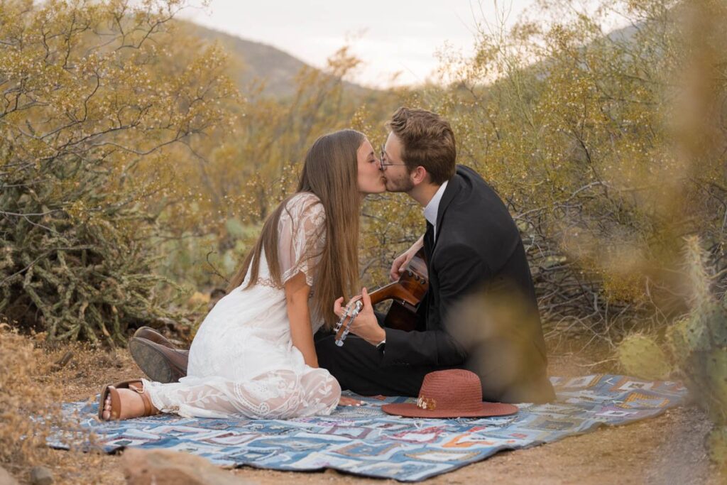 Bride and groom kiss sitting on blanket in desert while groom plays guitar.