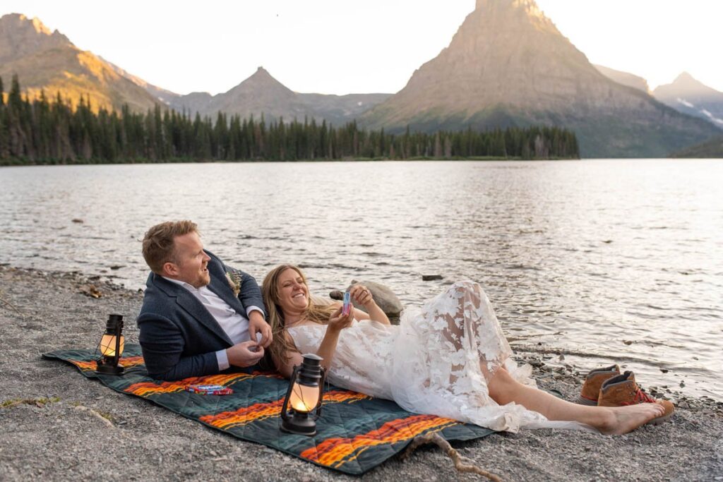 Bride and groom enjoy a candy picnic on a blanket near a lake in the mountains.