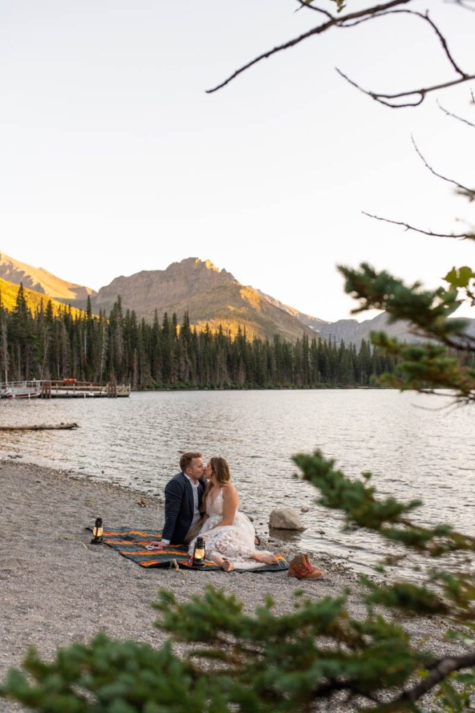 Couple kisses on blanket next a lake with mountains behind them.