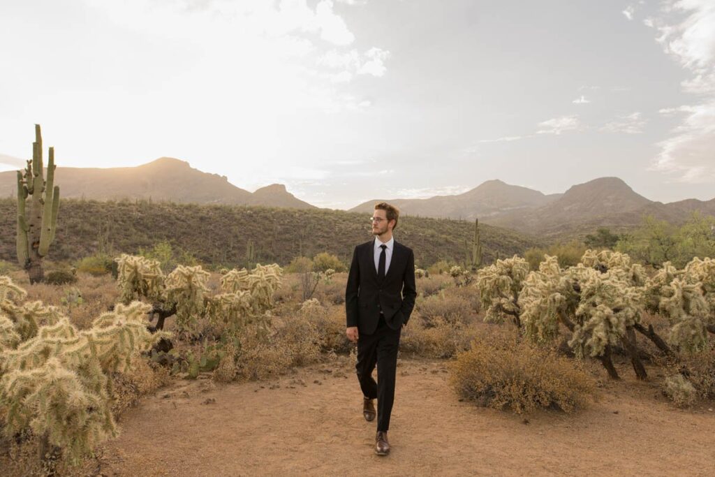 Groom walks with one hand in his pocket in the desert during sunset.