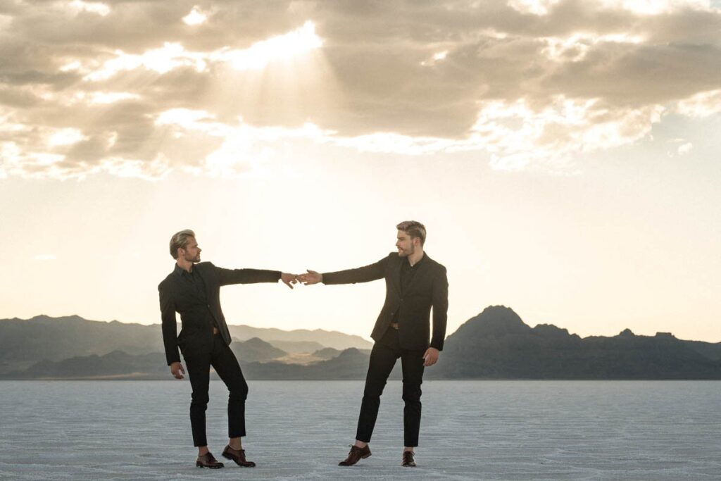 Grooms reach for each other's hands standing in the Salt Flats with sun rays behind them.