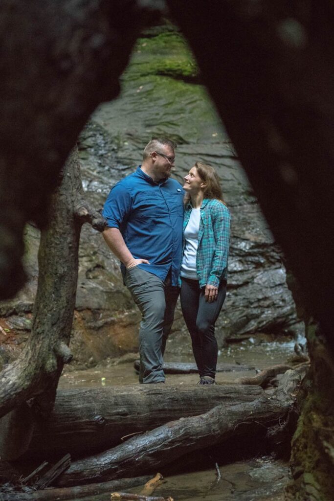 Couple stands between logs while looking happily at one another.