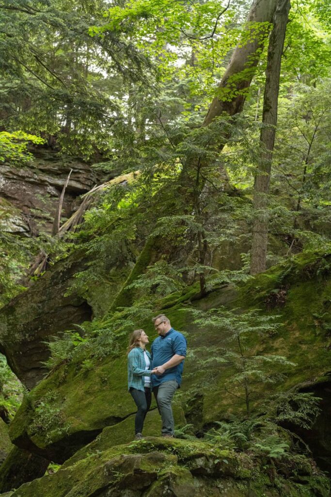 Couple looks at each other while standing on a mossy boulder.