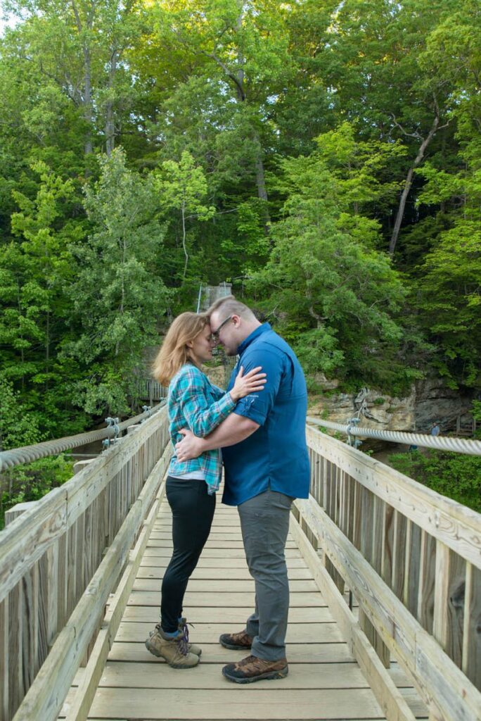 Couple rests foreheads together while standing on suspension bridge.