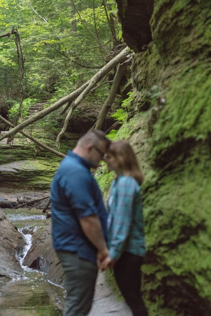 Blurred couple rests heads together with stream behind them in focus on a hike at Turkey Run State Park in Indiana.
