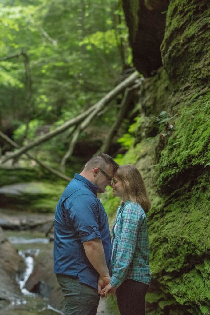 Couple holds hands while resting foreheads together in front of small stream.