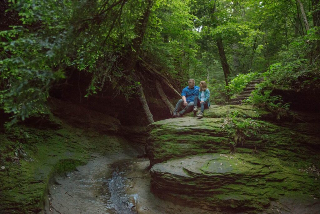 Couple sits on a large rock together with trees around them.