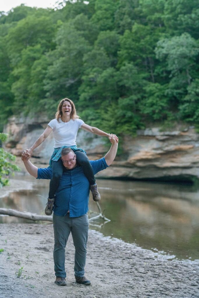 Woman laughs while man has her on his shoulders next to creek.