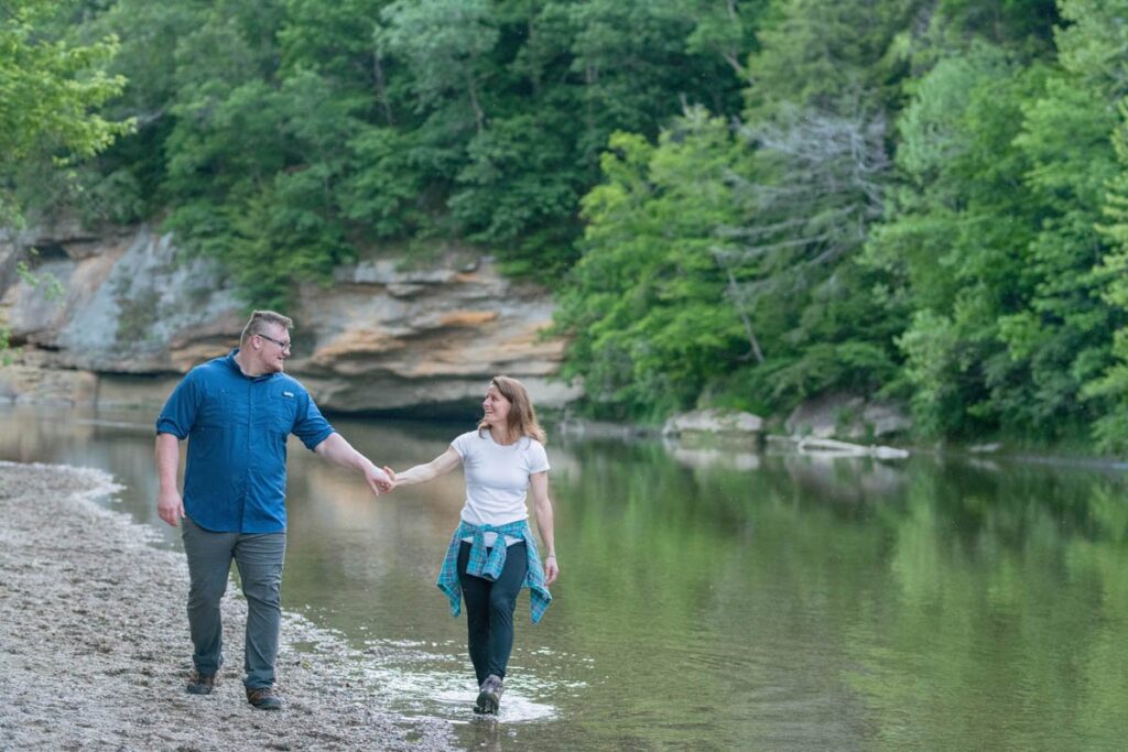 Couple looks at one another and smile while holding hands and walking in a creek.