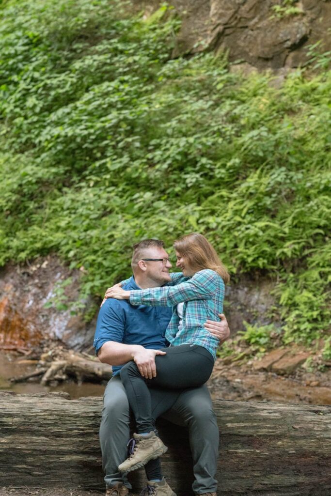 Man and woman laughing and sitting on a fallen log with green plants behind them.