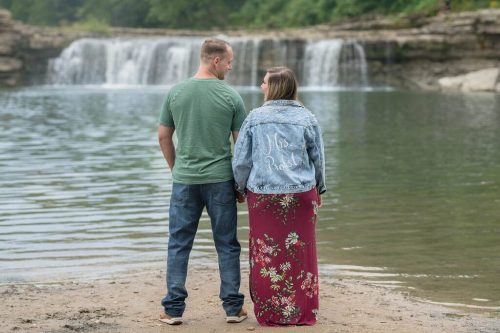 Couple looks at each other happily for their waterfall engagement photos with Cataract Falls in the background.