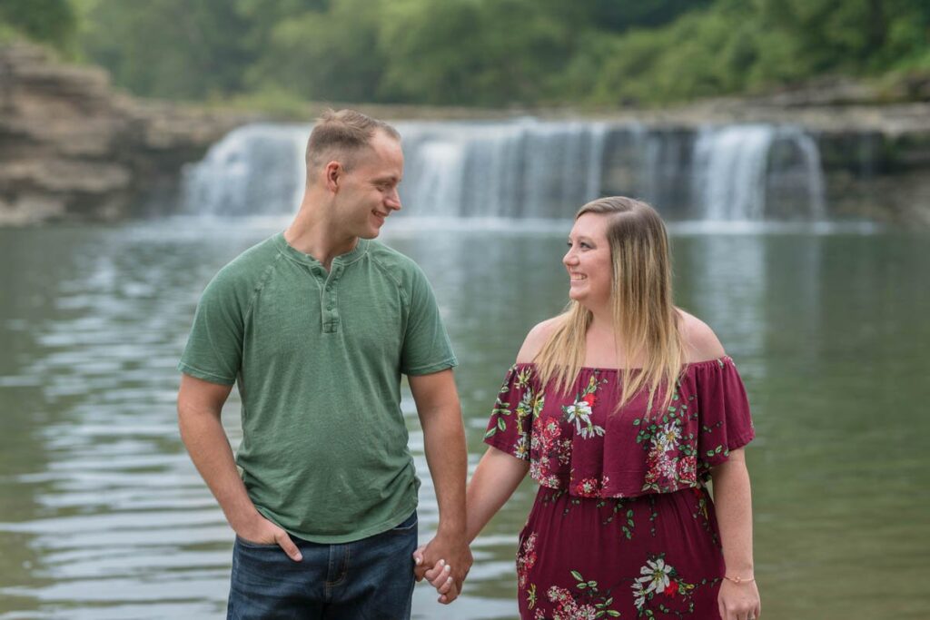 Couple smiles while looking at each other during their waterfall engagement photos with Cataract Falls behind them.