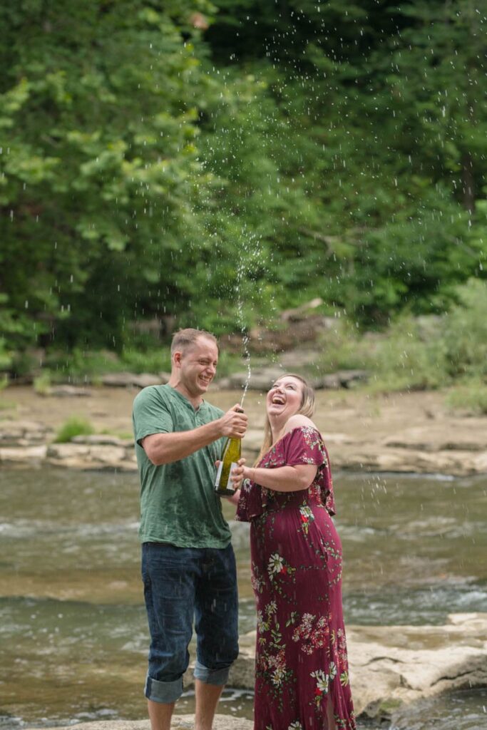 Couple laughs during a sparkling water pop during their waterfall engagement photos at Cataract Falls.