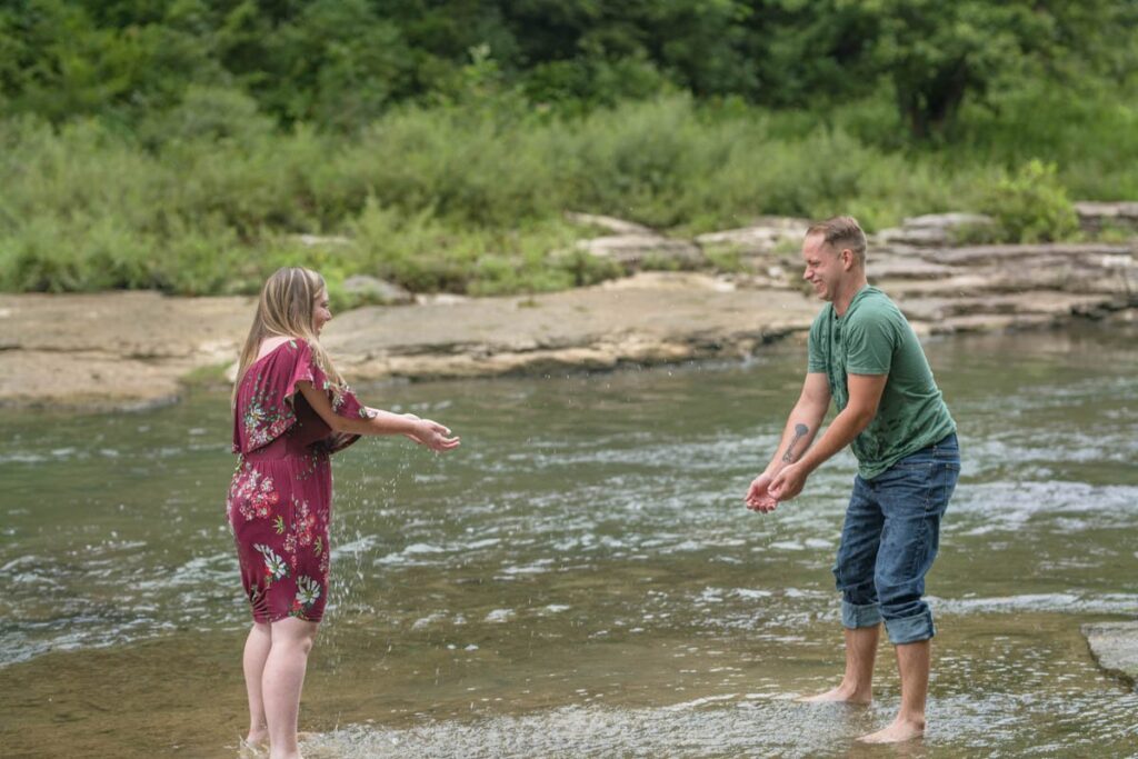 Couple tosses water at each other while laughing during their waterfall engagement photos.