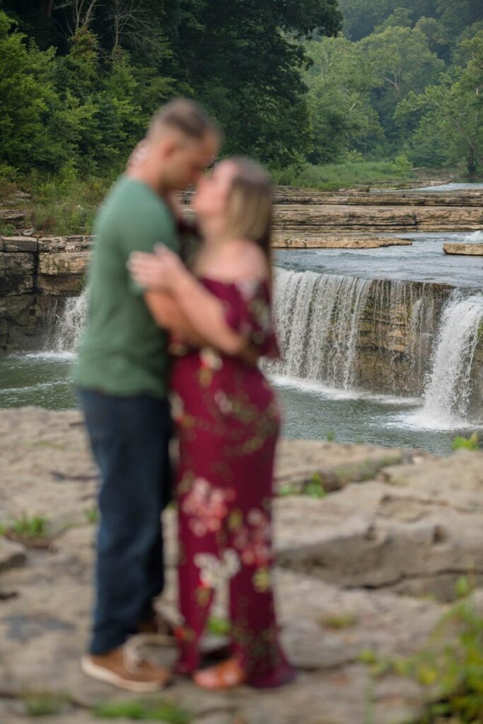 Blurry couple stands in front of in-focus Cataract Falls during their waterfall engagement photos..