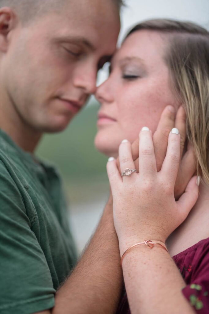Man holds woman's face closely and she rests her hand on his to show her engagement ring.