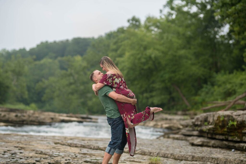 Man lifts woman up near water as they look at each other.
