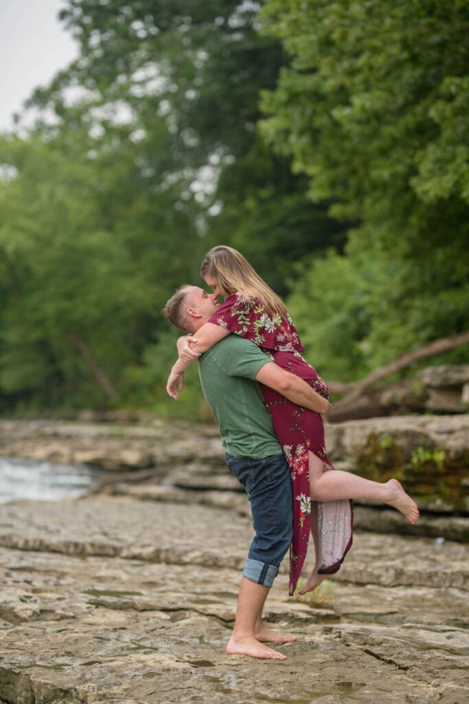 Man lifts woman up near water and they almost kiss.