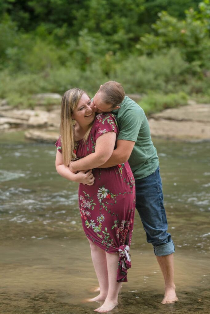 Man wraps his arms around woman and kisses her cheek while they stand in a stream.