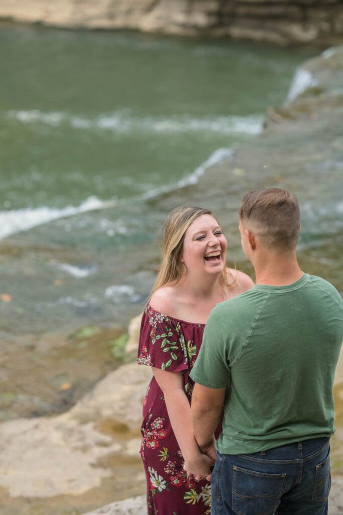 Woman is laughing looking at man during their waterfall engagement photos.