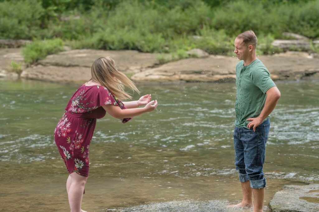 Woman splashes man with water in stream while laughing.