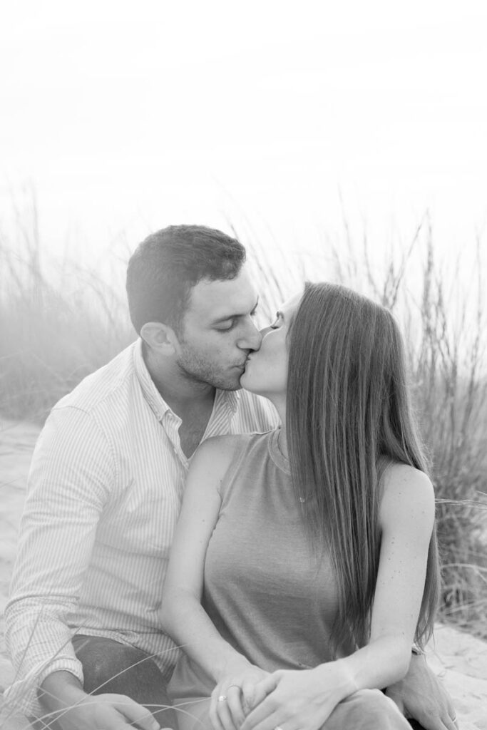 Couple kisses while sitting in tall beach grass.