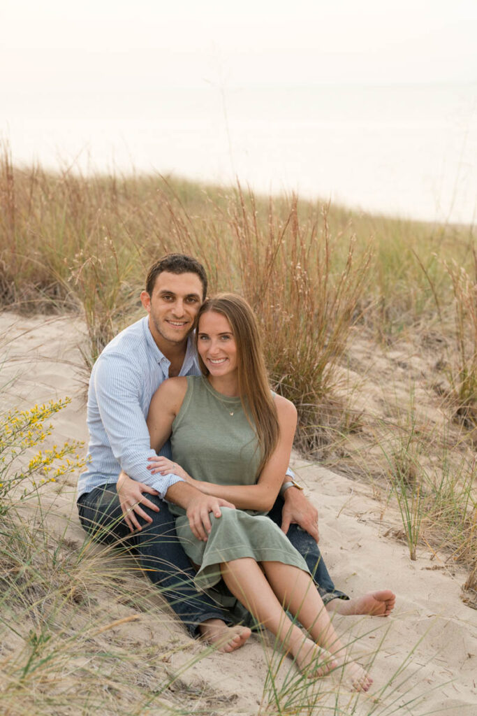 Couple sits in sand among beach grass smiling.
