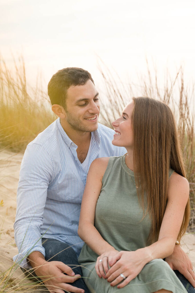 Couple looks at one another as they sit in the sand next to beach grass with the sunset behind them.