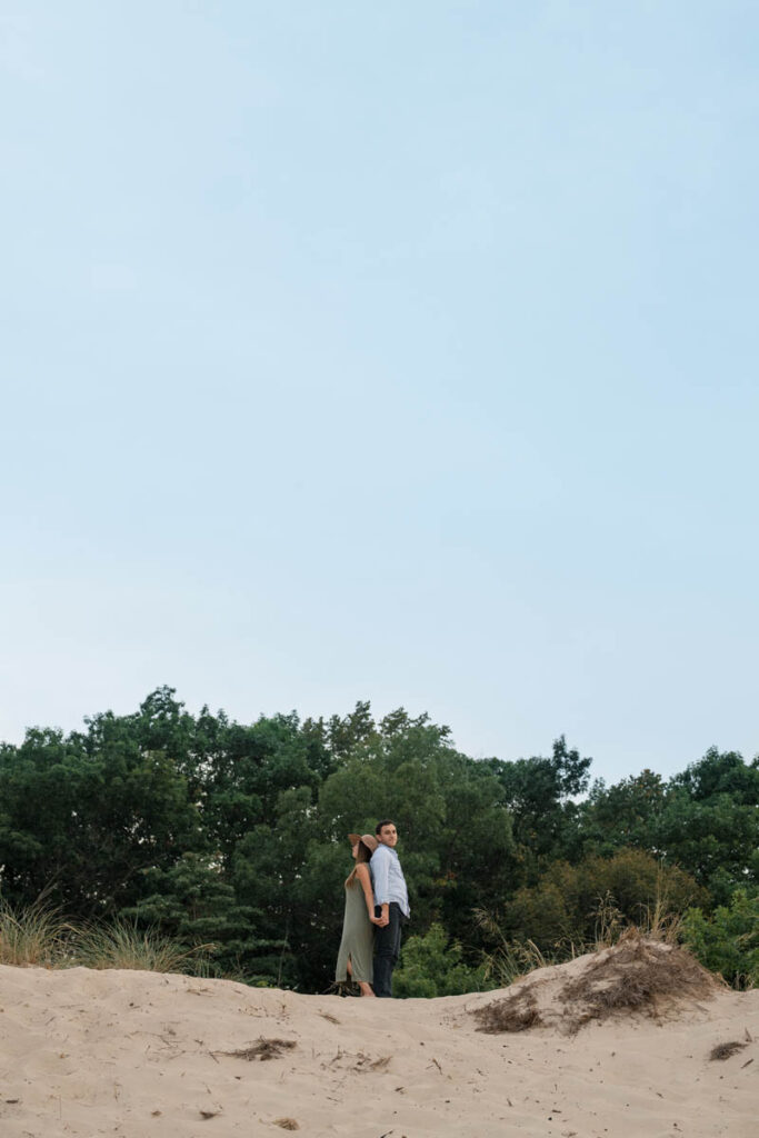 Couple stand on top of sand dune back to back.