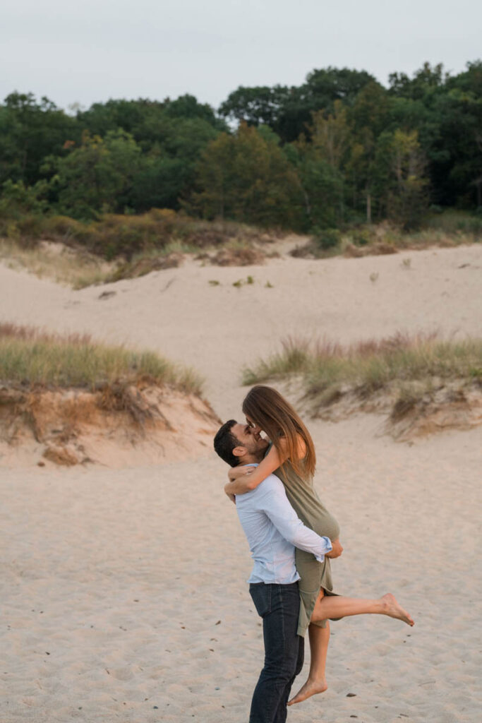 Man lifts woman and they get ready to kiss with big sand dunes behind them.