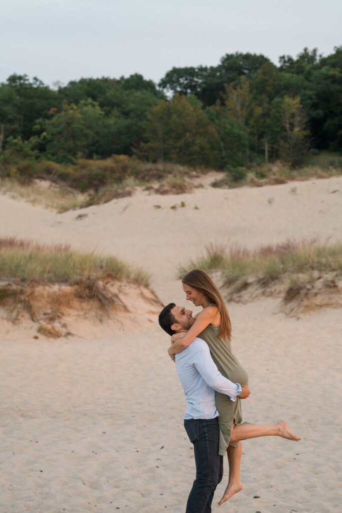 Man and woman laugh while he lifts woman up with sand dunes behind them.