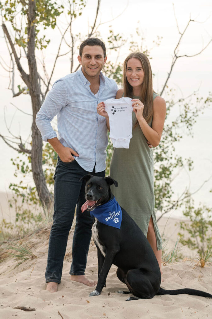 Couple smiles with their dog while holding a onesie for a pregnancy announcement.