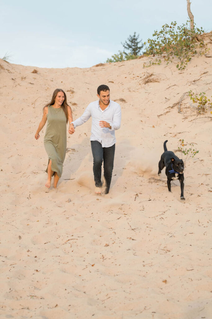 Couple is laughing while running down a sand dune with their dog.