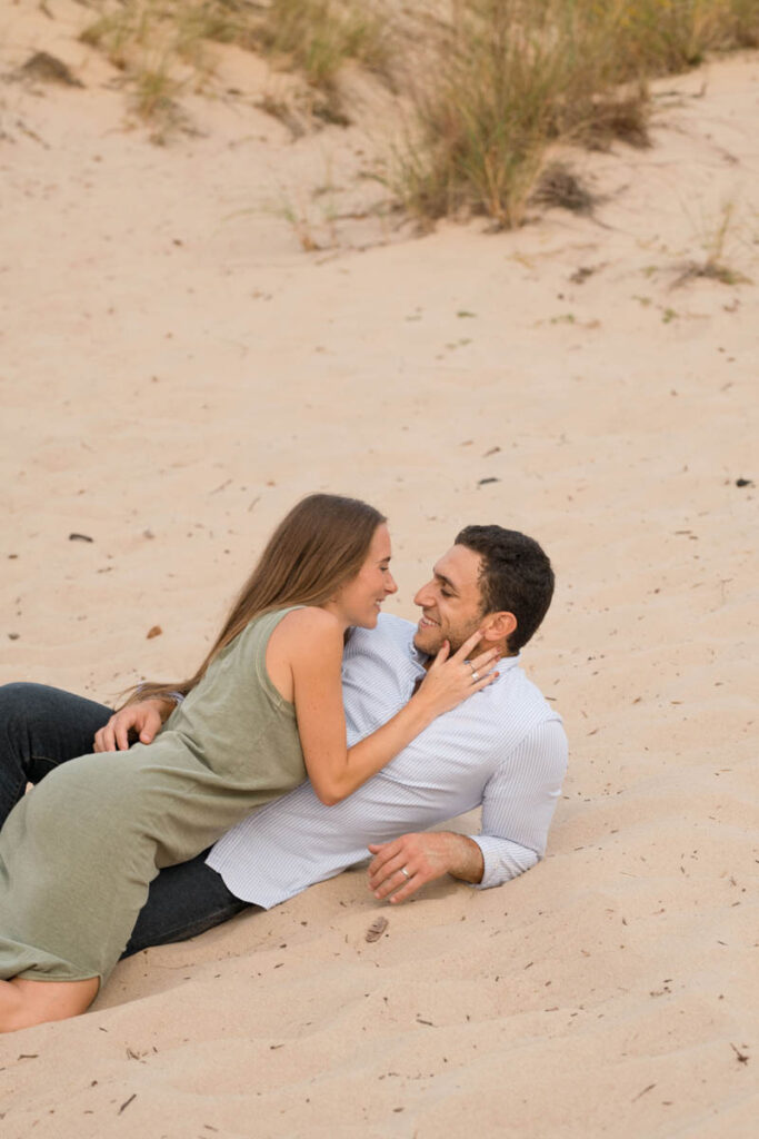 Woman holds man's face in her hand while they lay in the sand together.
