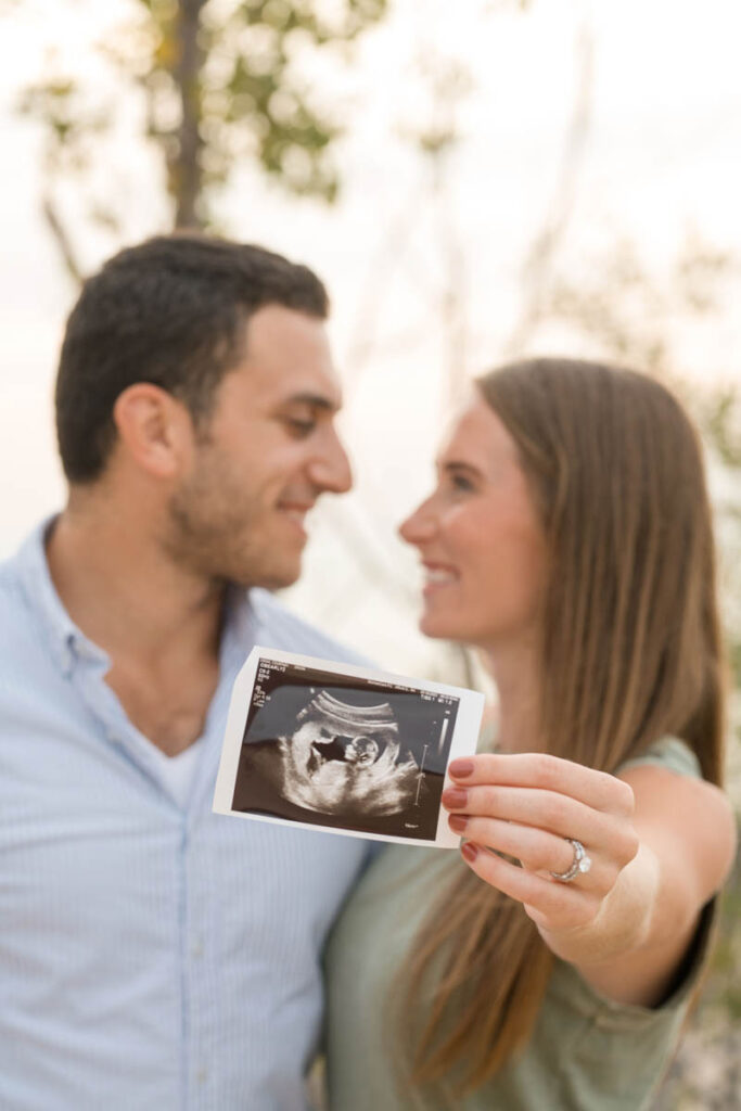 Couple holding up a sonogram photo and smiling at each other during beach couples photography session.