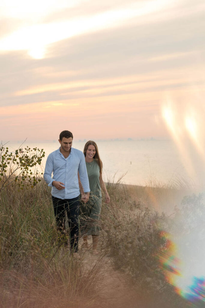 Couple walks through small sandy path among beach grass while sun sets behind them.