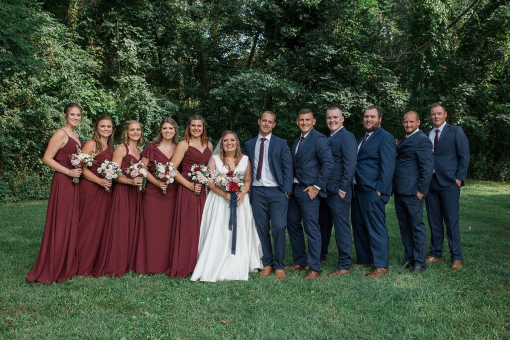 Bride and groom smile for portrait with their entire bridal party wearing navy and crimson.