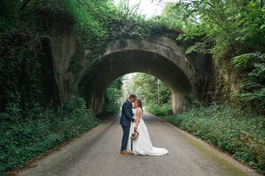Bride and groom stand with arms around each other in front of an old bridge.