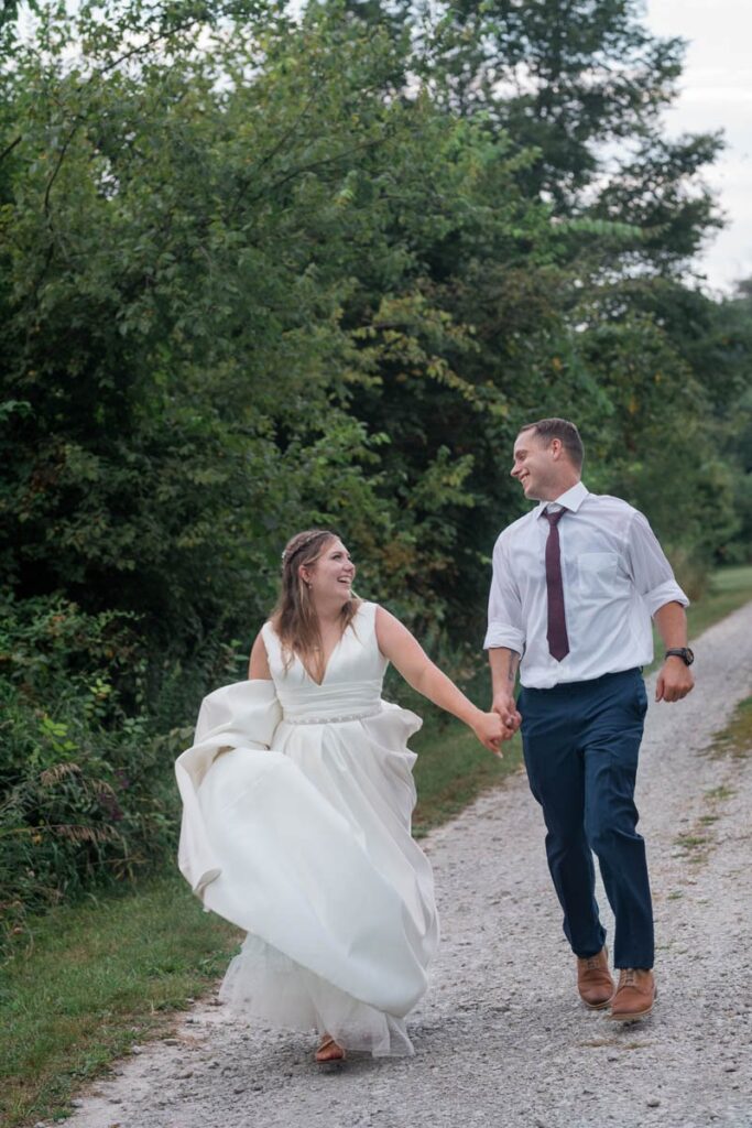 Bride and groom look at each other as they run holding hands down a gravel road.