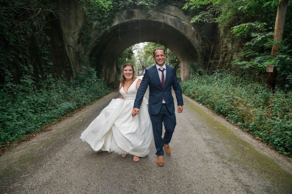 Bride and groom hold hands while walking away from an old bridge.