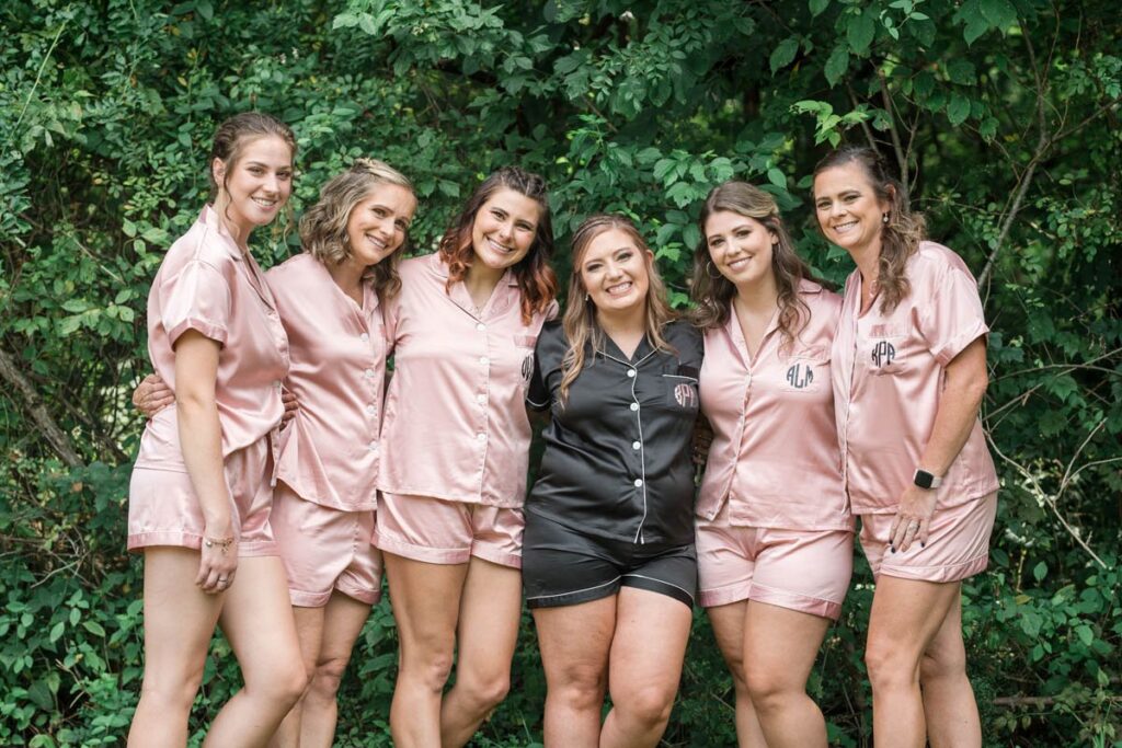 Bride and bridesmaids smile while wearing matching pink and black pajamas with monogrammed initials on them.