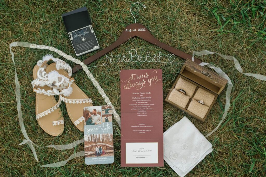 Flat lay image of wedding items including rings, shoes, jewelry, and invitations in the grass.